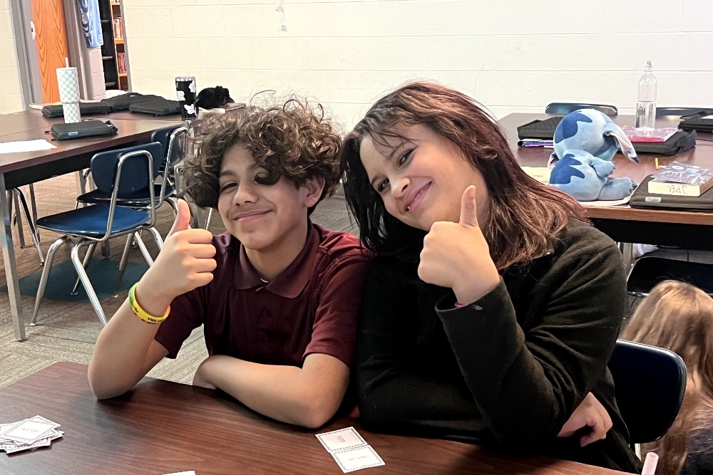 Two JPEC students are sitting together in a classroom and giving a thumbs up.